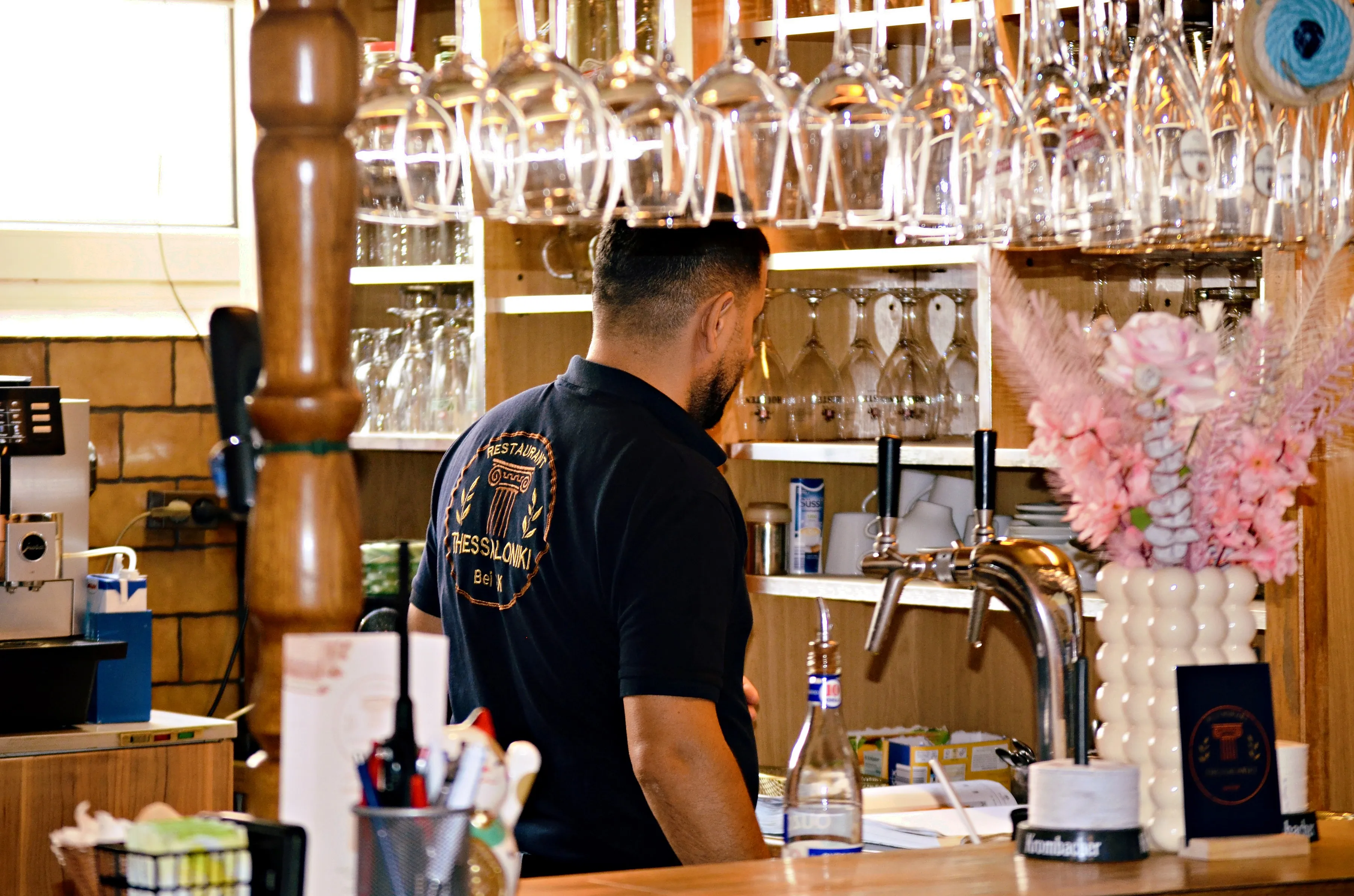 Kristo hinter der Bar, darüber hängende Weingläser